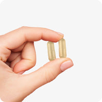 Close-up of a hand holding two beige Conceive Plus Prenatal vegan capsules against a white background, highlighting capsule size and texture for prenatal supplement use.