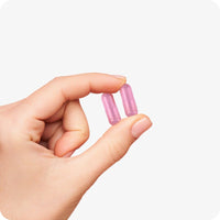 Hand holding two pink Conceive Plus Women's Fertility Support capsules between thumb and index finger on a white background, showing capsule size and color.