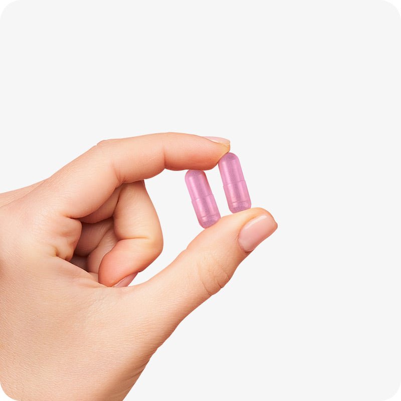 Hand holding two pink Conceive Plus Women's Fertility Support capsules between thumb and index finger on a white background, showing capsule size and color.